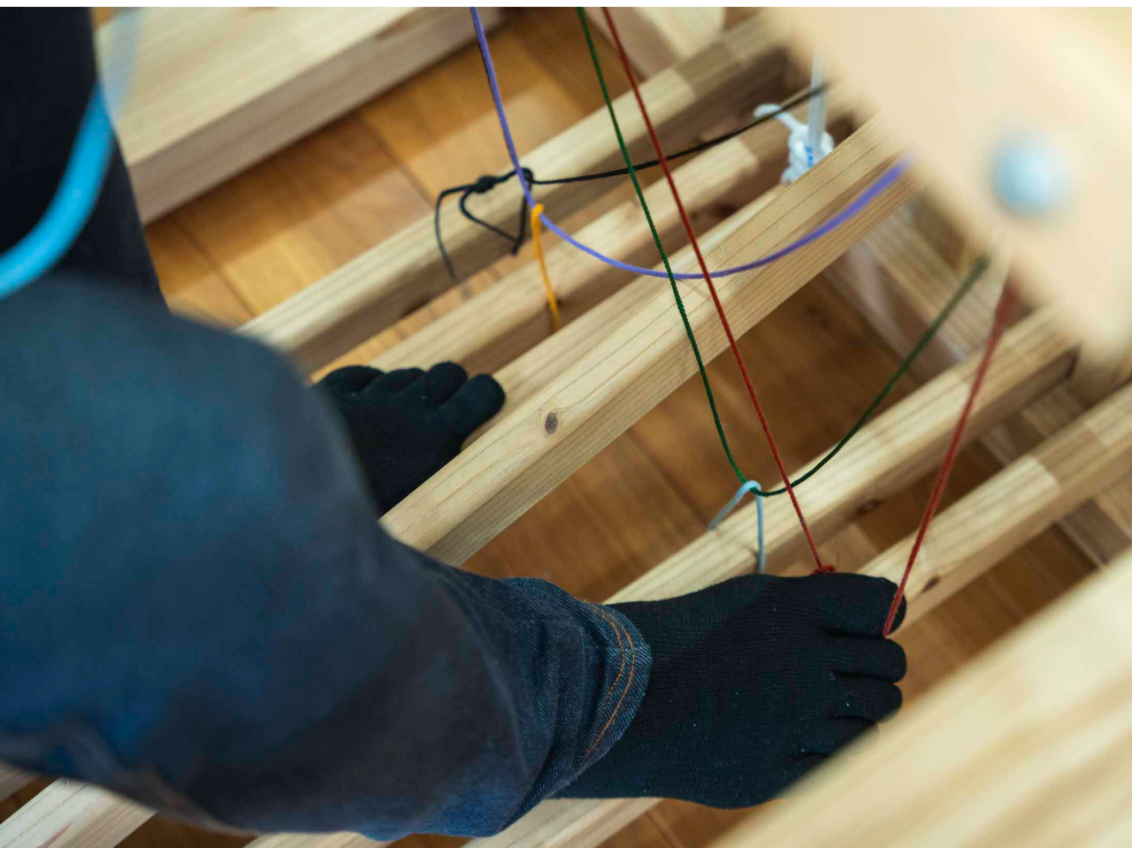 足元の踏み木(ペダルのような板)で本綜絖(ほんそうこう)を操作し、ヒモを足でひっぱって花綜絖(はなそうこう)を操作します。