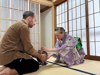 A guest is learning the art of handling a tea jar with guidance from the host.